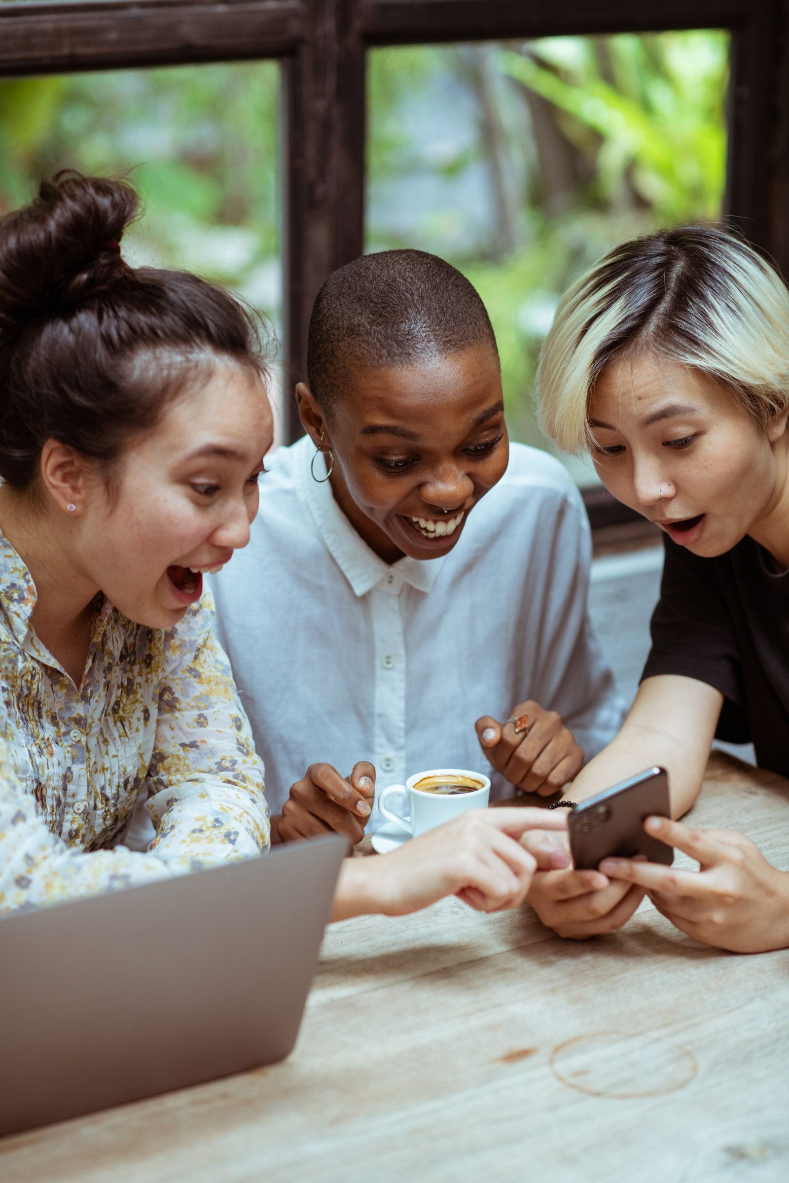 Three friends huddled around one phone at a café table, delighted